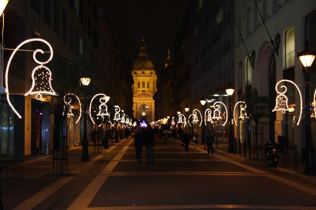 A Budapest street at night with Christmas lights shaped like bells and the lit dome of St Stephen's Basilica at the far end.